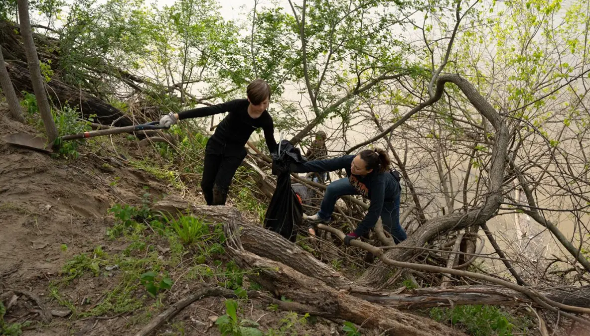Two people climbing a steep river embankment carrying garbage bags while cleaning the river